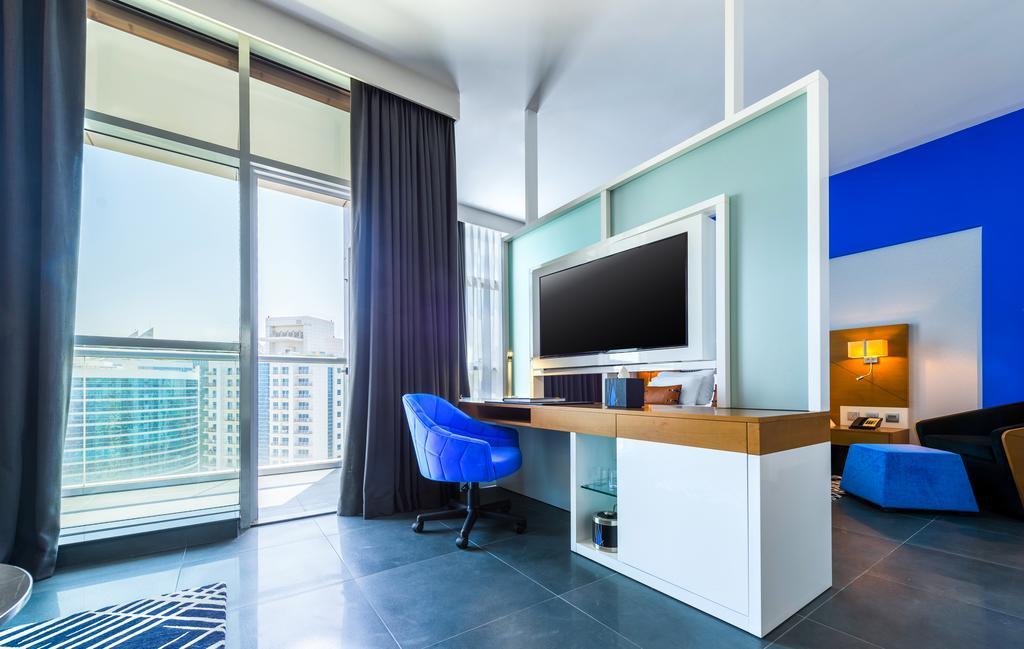 Hotel Tryp By Wyndham Dubai Esterno foto The photo shows a modern room with large windows allowing natural light to enter. There's a stylish desk area featuring a flat-screen TV mounted on the wall, along with a light wooden desk and a blue chair. The wall behind the TV is painted in a ligh